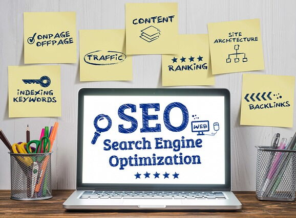 Laptop auf einem Tisch. Auf dem Bildschirm steht SEO Search Engine Optimization. An der Wand über dem Laptop kleben Post-Its mit INDEXING KEYWORDS, ONPAGE/OFFPAGE,TRAFFIC, CONTENT, RANKING, SITE ARCHITECTURE und BACKLINKS