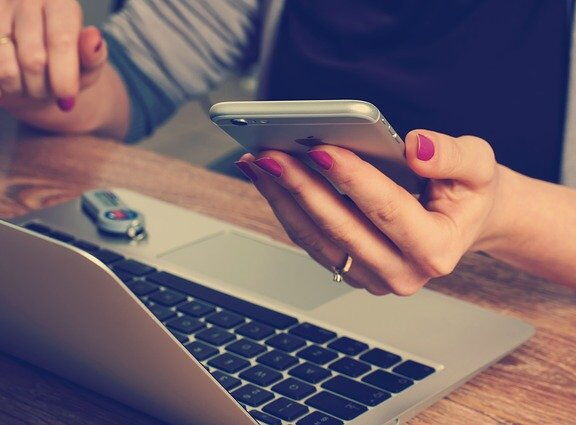 Eine Frau am Laptop mit einem Smartphone in der Hand. der Kopf ist abgeschnitten.