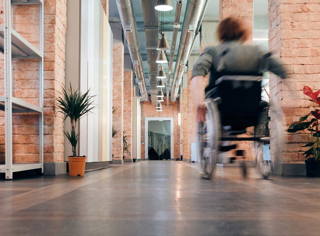 Verschwommene weibliche Person fährt mit Rollstuhl einen Gang in einem Bürogebäude aus rotem Backstein entlang vom Betrachter weg..